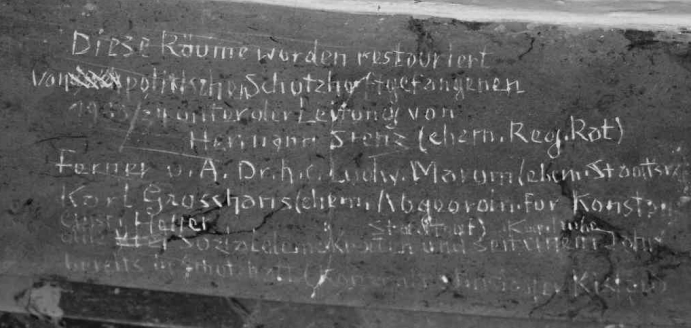 Weißer handgeschriebener deutscher Text ist auf einer dunklen, rauen Wandoberfläche eingeschrieben. Die Schrift ist ungleichmäßig und teilweise verblasst, was auf das Alter hinweist. Einige Textzeilen sind klar, während andere schwieriger zu entziffern sind.