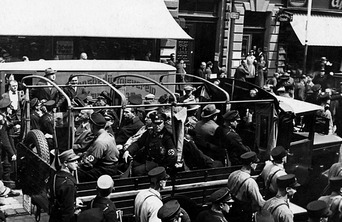 Ein Schwarz-Weiß-Foto, das Nazi-Offiziere und Männer in Uniform in einem offenen Lastwagen während einer belebten Straßenszene in Deutschland zeigt, mit Schaulustigen in der Nähe und Gebäuden im Hintergrund.