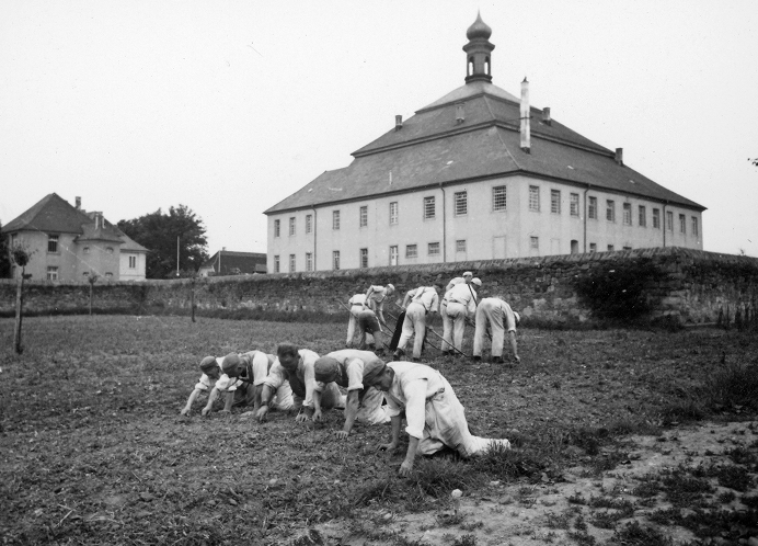Eine Gruppe von Menschen in Uniform kriecht und arbeitet an einem bedeckten Tag auf einem Feld in der Nähe eines großen, historischen Gebäudes mit kuppelförmigem Dach und Turm, das von einer Steinmauer umgeben ist.