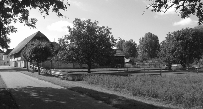 Ein Schwarz-Weiß-Foto von einer friedlichen ländlichen Szene mit einer großen Scheune, einem Bauernhaus, eingezäunten Flächen, Bäumen und einer ruhigen Straße, die an den Gebäuden vorbeiführt, unter einem teilweise bewölkten Himmel.