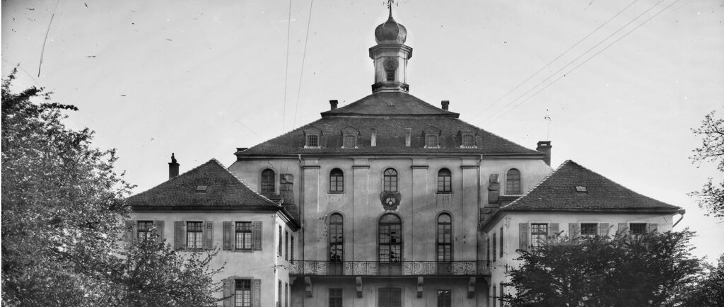 Schwarz-Weiß-Foto eines großen, historischen Gebäudes mit einem zentralen Turm und einer Kuppel, umgeben von Bäumen. Das Gebäude hat hohe Fenster, einen Balkon und ein steiles, mit Ziegeln gedecktes Dach.