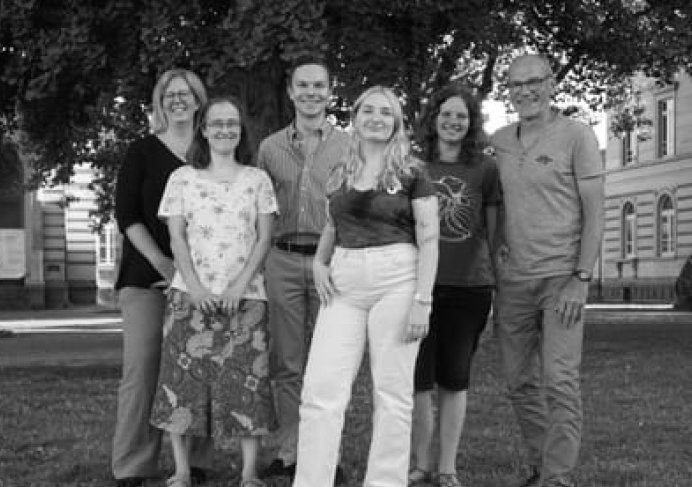 Sechs Erwachsene stehen zusammen auf einer Wiese und lächeln in die Kamera. Im Hintergrund sind Bäume und Gebäude zu sehen. Das Foto ist in schwarz-weiß gehaltenen . 